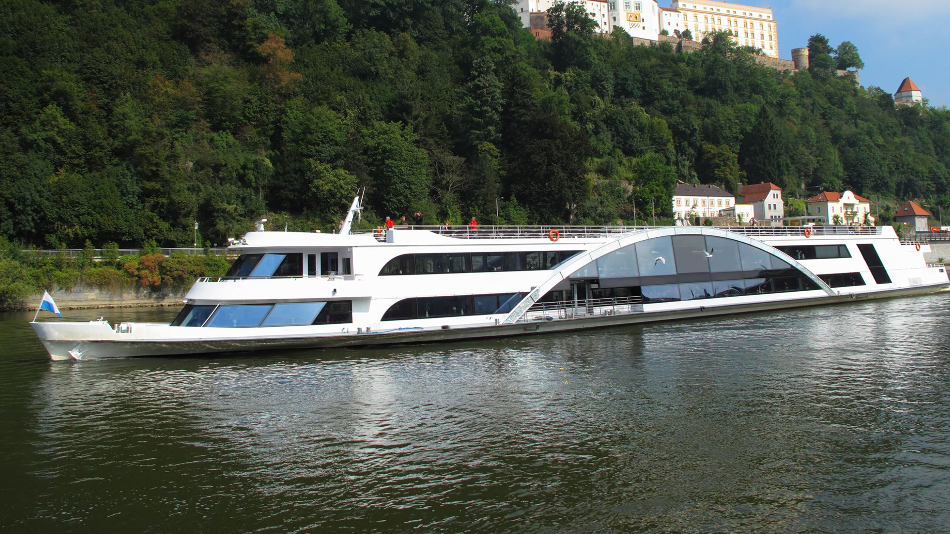 Sleek white river boat with a curved glass deck below a wooded hillside