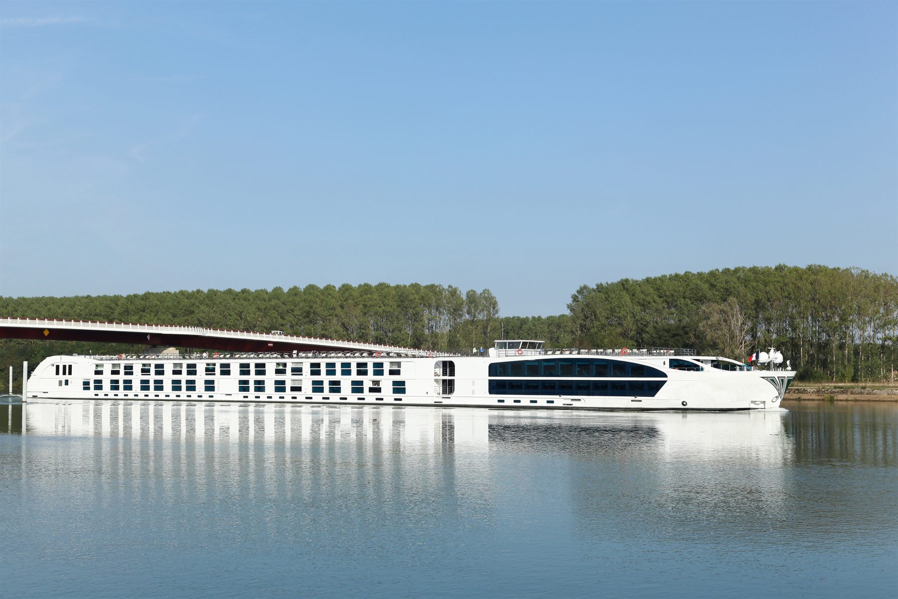 Modern white river cruise ship reflected in calm water beside a tree-lined bank
